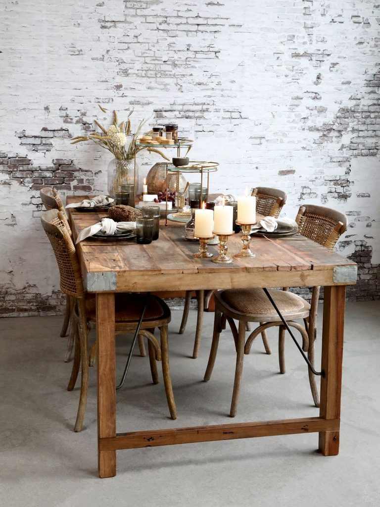 Amelie dining range with rustic wooden table and woven chairs, styled with candles and tableware against a whitewashed brick wall.